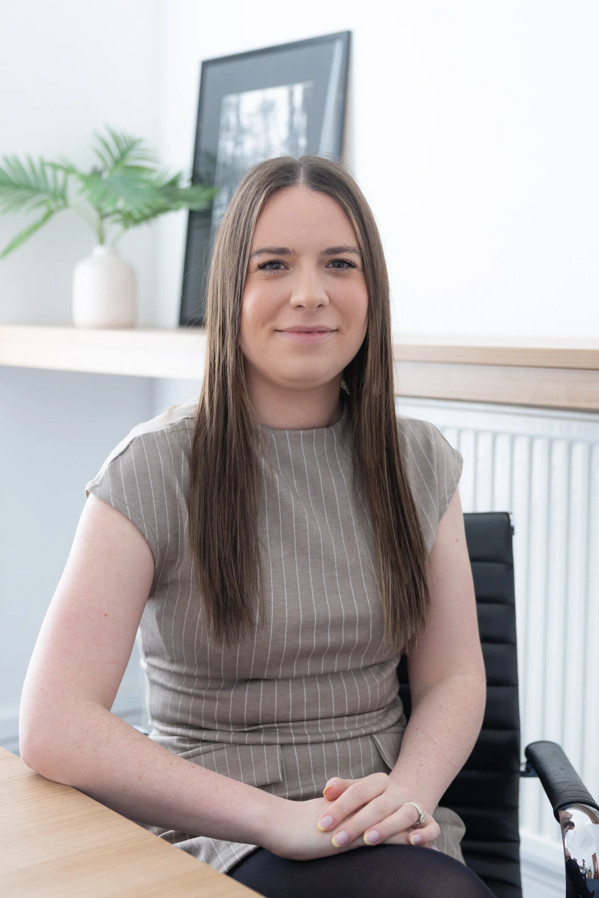 Woman with long brown hair sits at a desk in a bright office, wearing a beige striped top, hands folded on her lap.