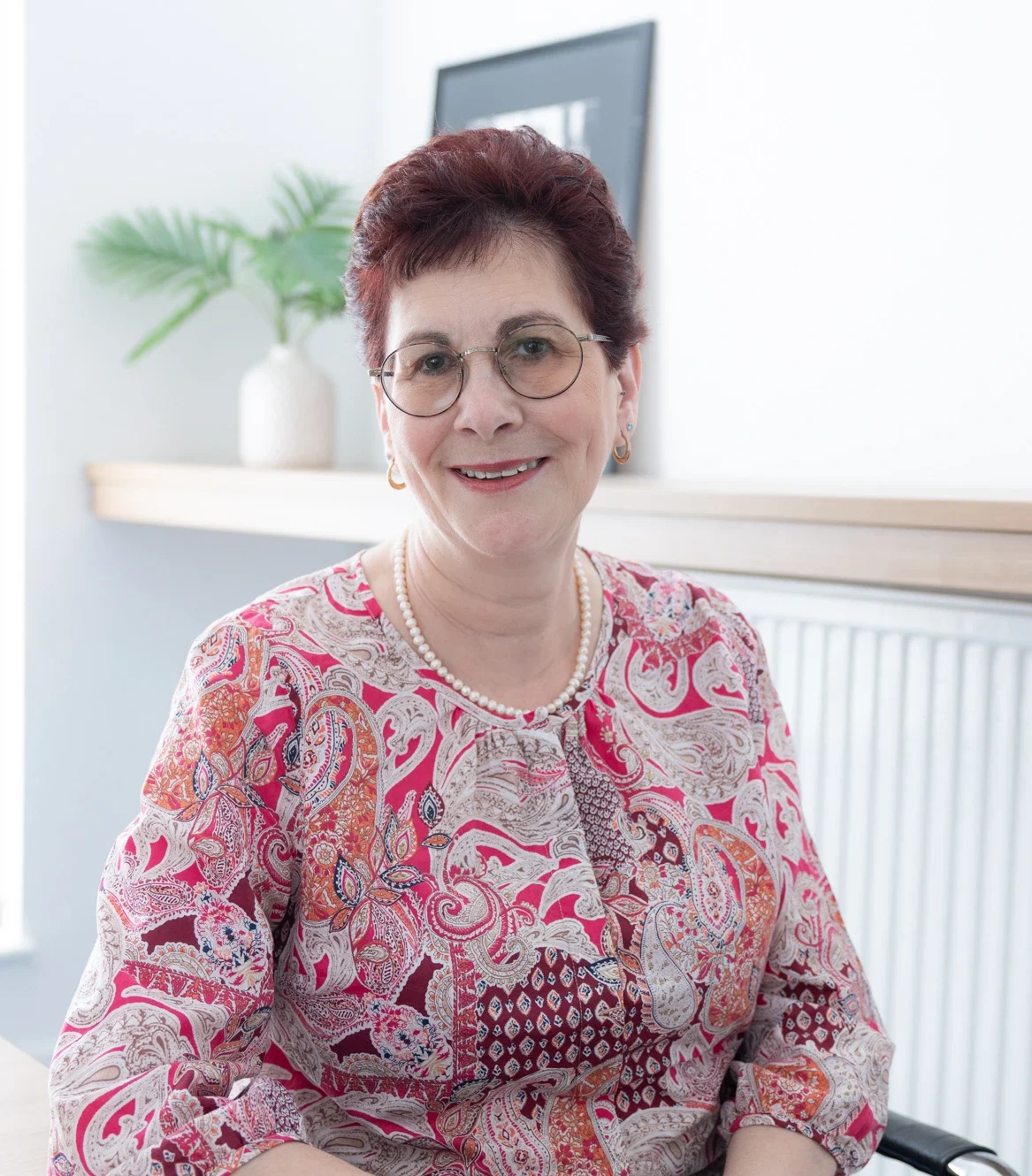 Smiling older woman with short burgundy hair and round glasses, wearing a pink paisley blouse and pearl necklace, seated at a desk.