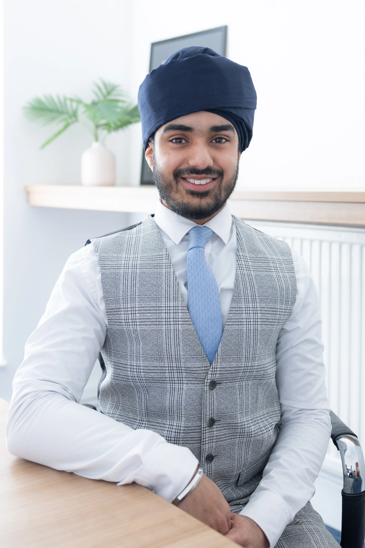 Professional man wearing a navy turban, gray plaid vest, white shirt, and light blue tie sits at a desk in a bright office setting, smiling at the camera.
